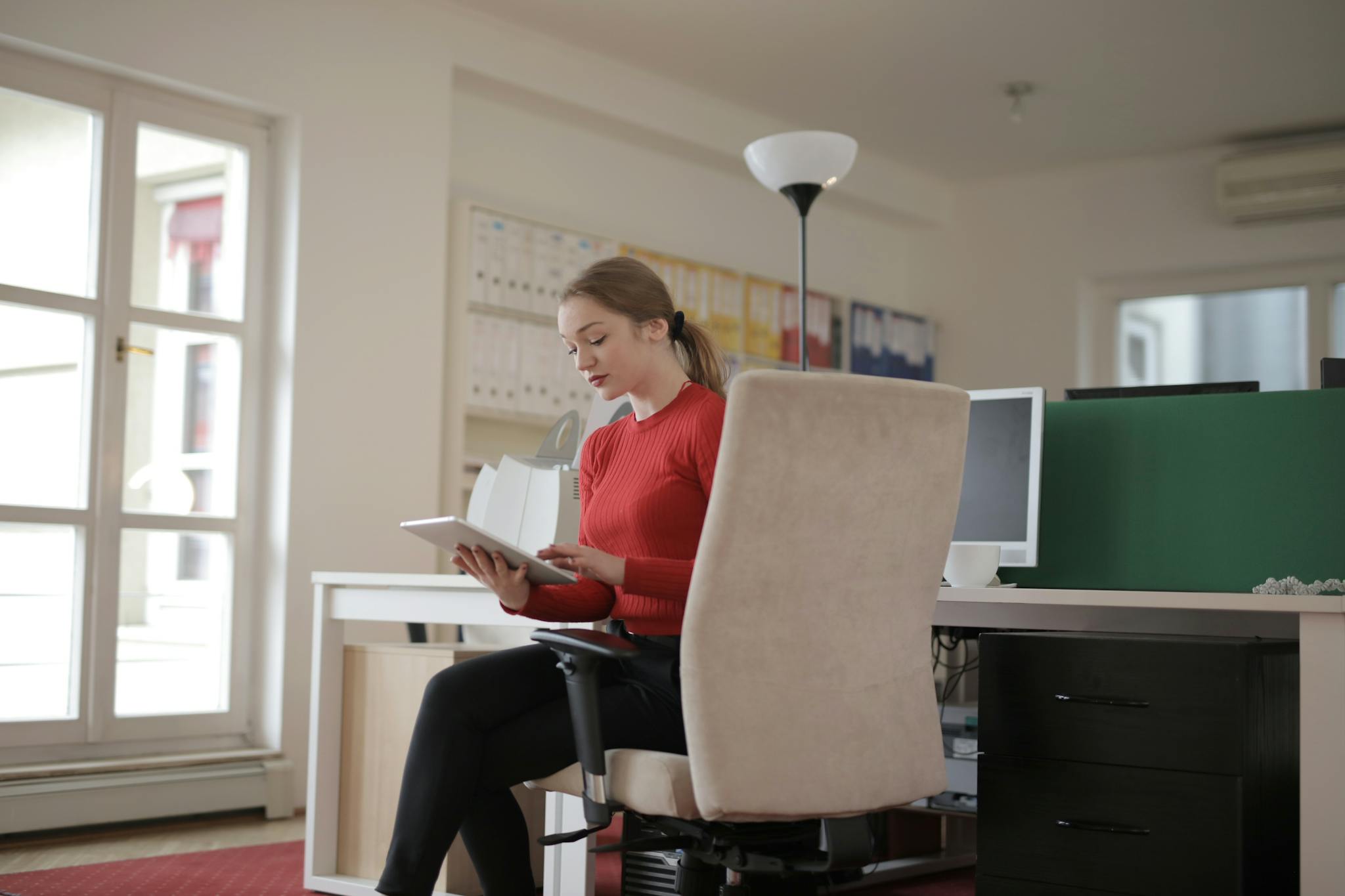 Young woman in a red sweater working on a tablet in a bright office environment.